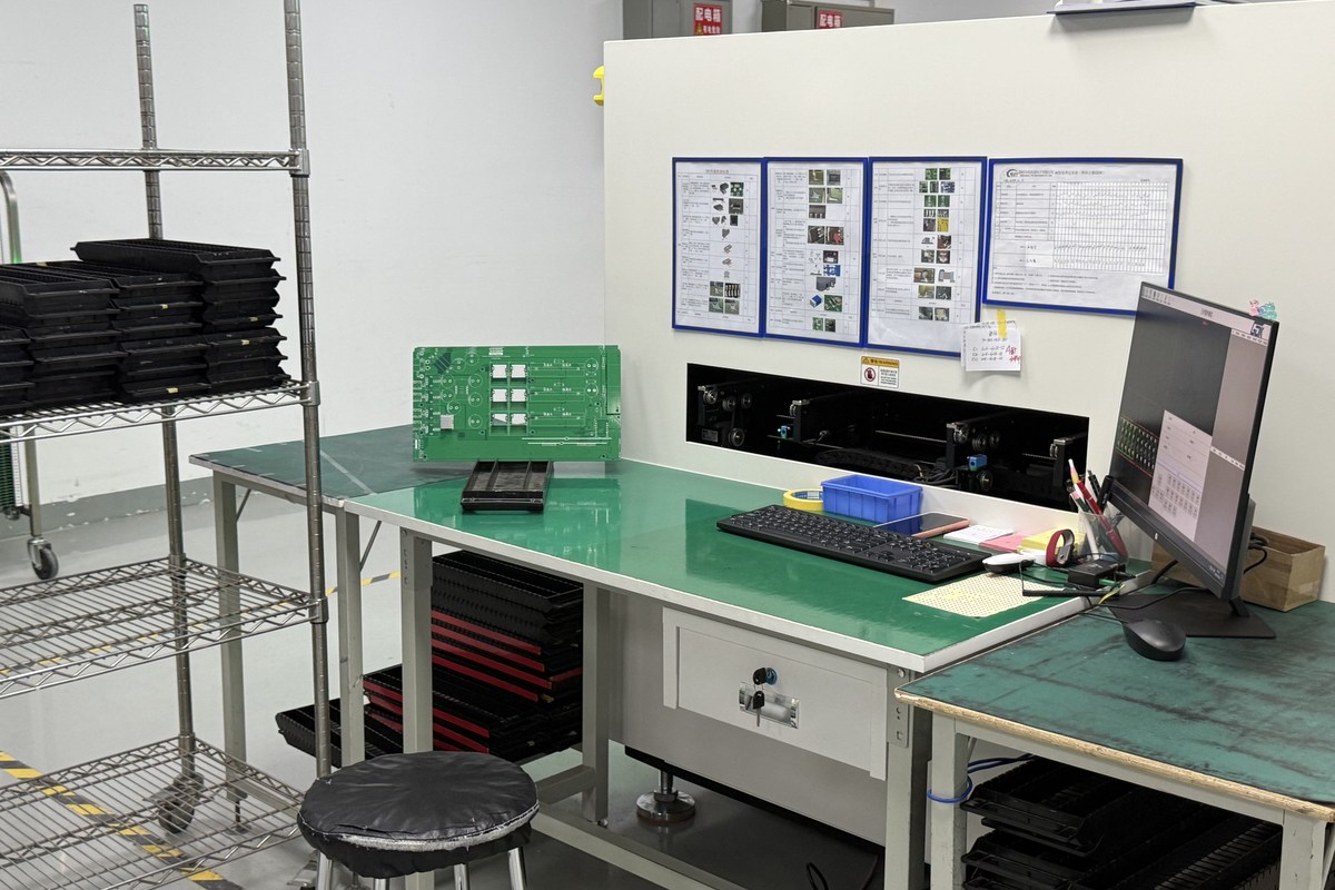 Real factory photo of a bare green PCB beside a board inspection workstation and production trays
